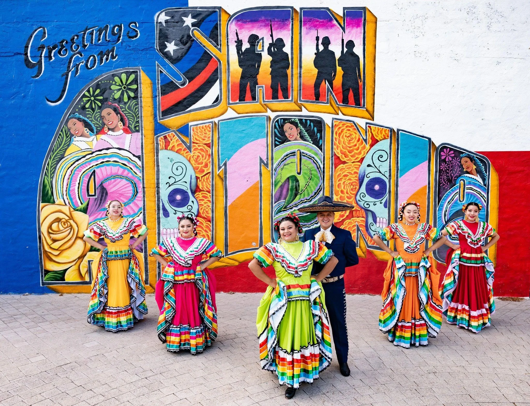Grupo Folklórico de Bendiciones posing in colorful traditional attire in front of the Greetings from San Antonio mural